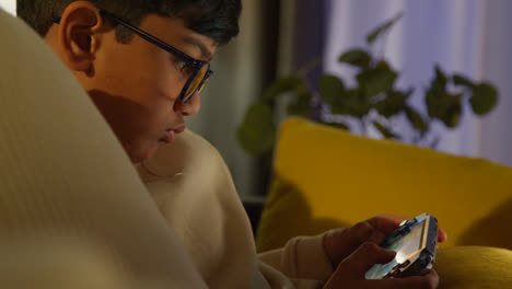 Young-Boy-Sitting-On-Sofa-At-Home-Playing-Game-Or-Streaming-Onto-Handheld-Gaming-Device-At-Night-3